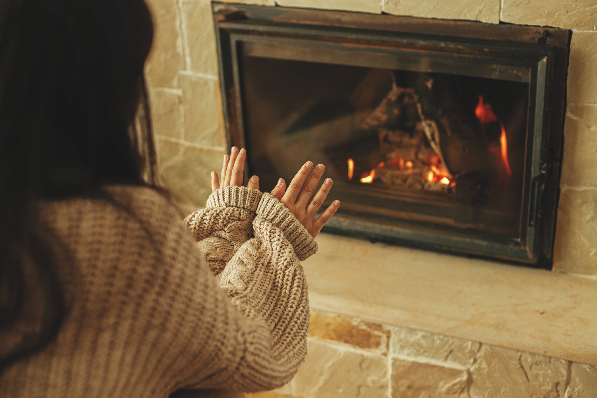 Woman in cozy sweater warming up hands at fireplace in rustic room. Fireplace heating in winter